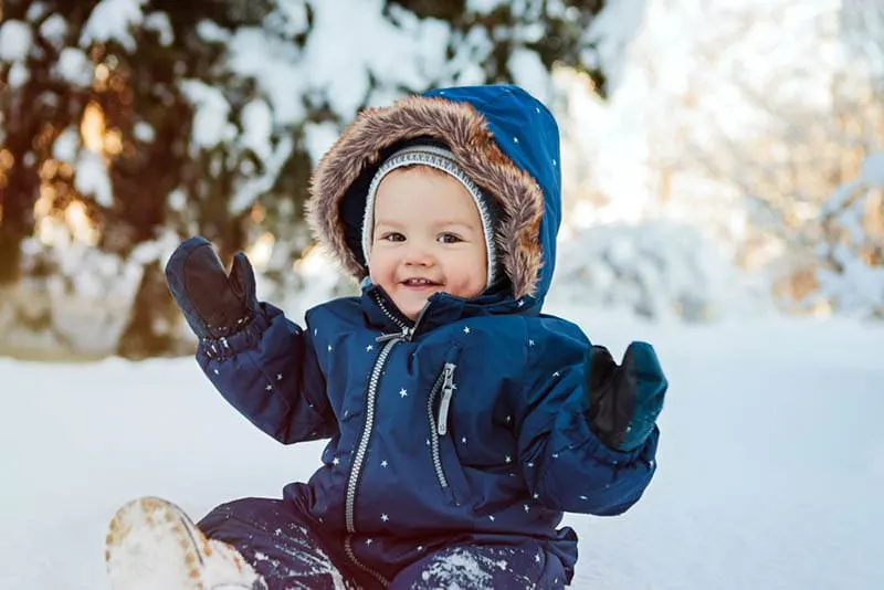 schöne Baby Junge trägt Winterkleidung und sitzt auf dem Schnee im Freien