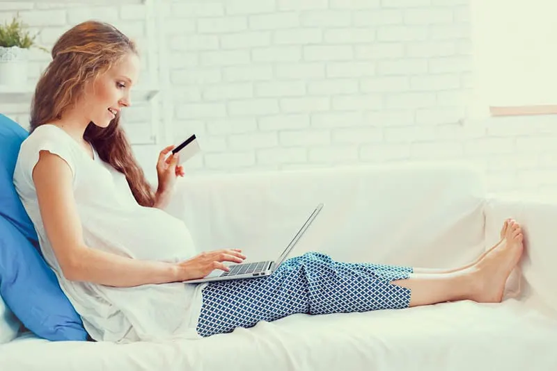 schöne schwangere Frau sitzt auf der Couch mit Laptop und eine Kreditkarte und tun ein Online-Shopping