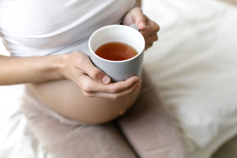schwangere Frau, die auf dem Bett sitzt und warme Tasse Tee hält