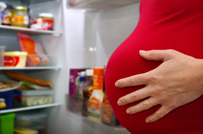 schwangere Frau hält Hand auf den Bauch und Blick auf Lebensmittel in geöffneten Kühlschrank