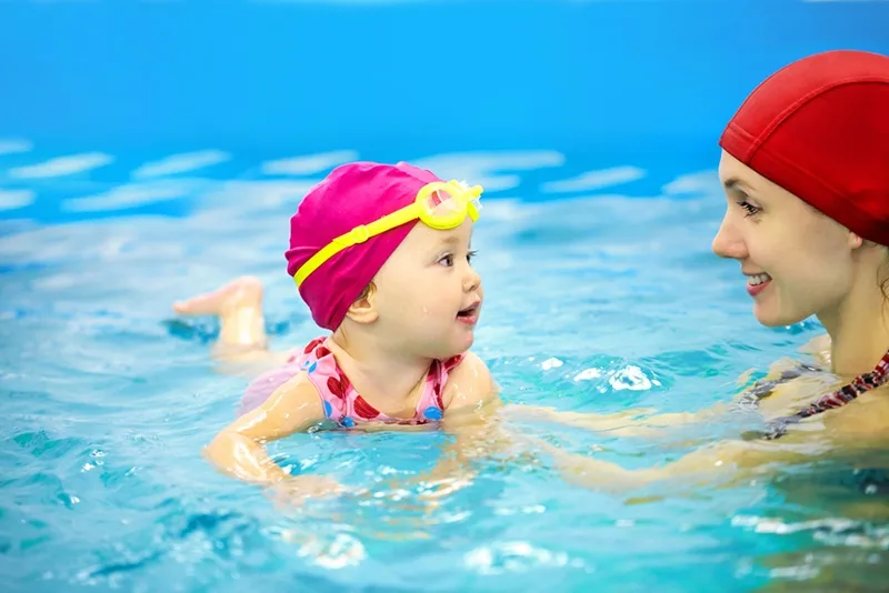 süßes Baby mit Schwimmbrille auf dem Kopf, der mit Mutter im Pool schwimmt