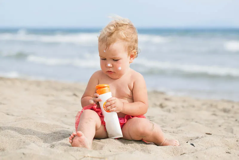 süßes kleines Baby, das mit Sonnencreme im Sand am Strand spielt