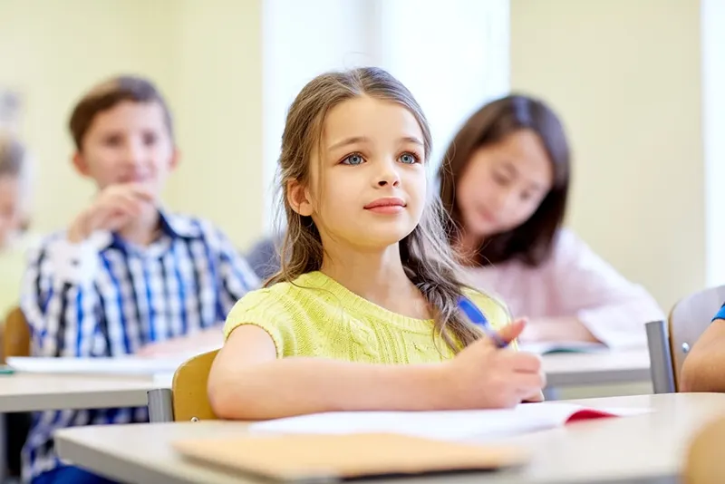süßes kleines Mädchen mit blauen Augen, das im Klassenzimmer mit Klassenkameraden sitzt