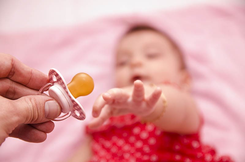 Baby-Mädchen liegt und versucht, Schnuller aus der Hand der Mutter zu fangen