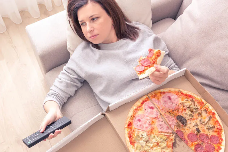 Junge Frau isst Pizza auf der Couch mit Pizzakarton und Fernbedienung