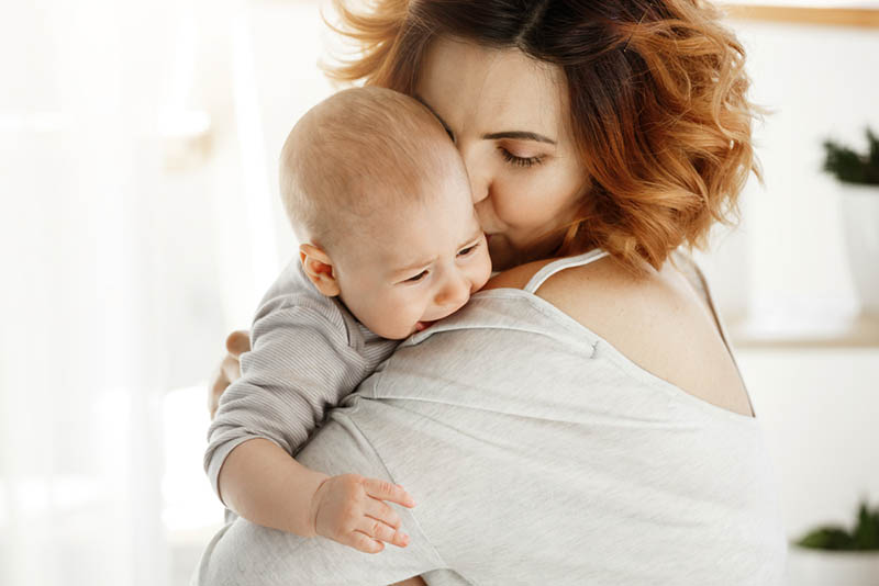 Junge Mutter umarmt und beruhigt ihr weinendes Baby zu Hause