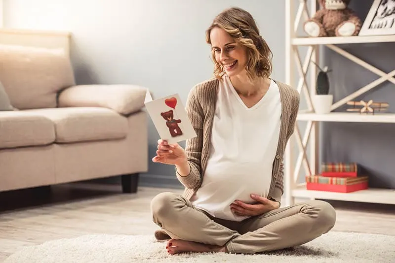 glückliche schwangere Frau, die auf dem Boden sitzt und eine Babykarte liest
