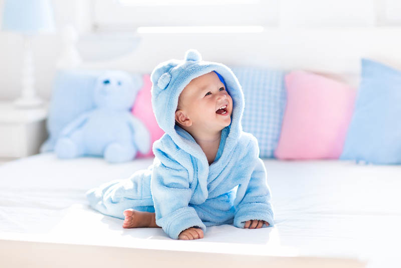 glückliches Baby Junge trägt blaue Dusche stricken und sitzt auf dem Bett