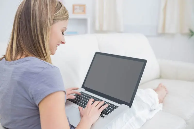 junge lächelnde Frau sitzt auf der Couch und tippt auf Laptop