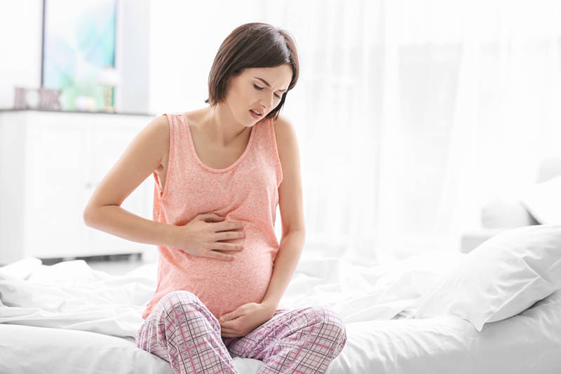 junge schwangere Frau hält für Bauch, während das Gefühl Schmerz auf dem Bett