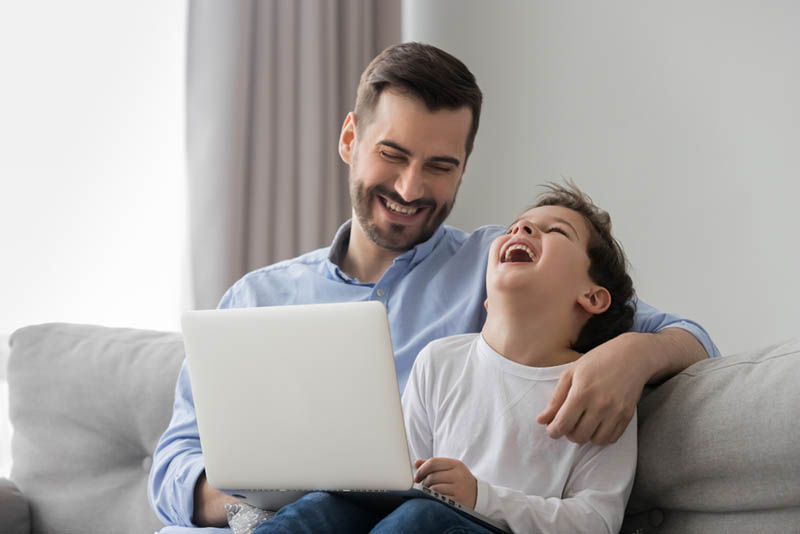 junger Vater sitzt auf der Couch mit laptopand lachend mit seinem Sohn 
