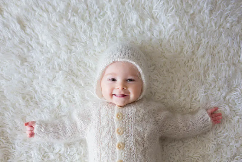 kleiner Baby-Junge in weißen gestrickten Strampler und eine Mütze lächelnd auf dem Bett