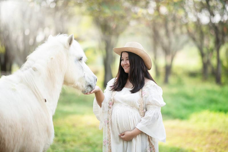 lächelnd schwangere Frau streichelt weißes Pferd im Freien