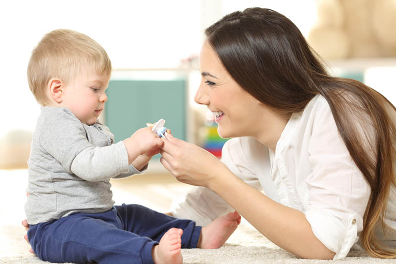 lächelnde Mutter gibt Schnuller zu ihrem Baby auf dem Boden