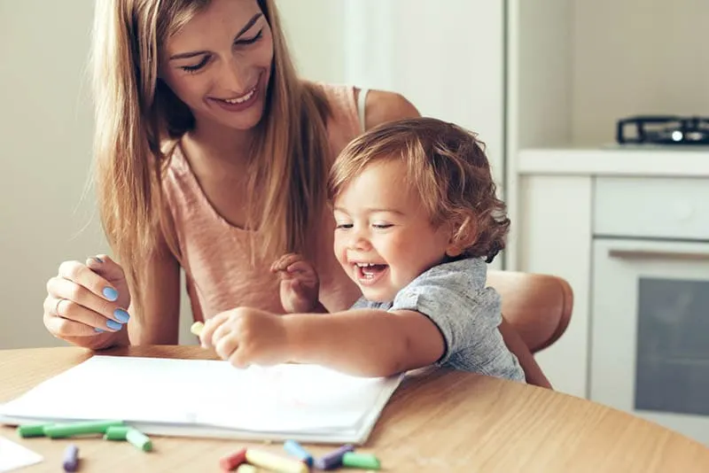 lächelnde Mutter hilft ihrem süßen Sohn beim Zeichnen am Tisch zu Hause