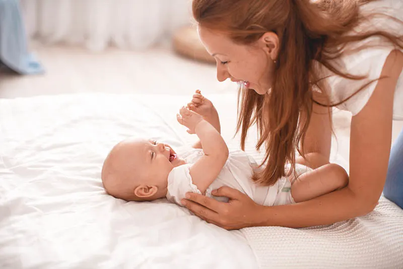 lächelnde Mutter spielt mit Baby auf dem Bett in weißen Laken