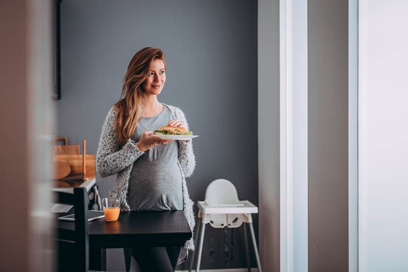 lächelnde schwangere Frau hält ein Sandwich auf Teller mit Glas Saft auf dem Tisch