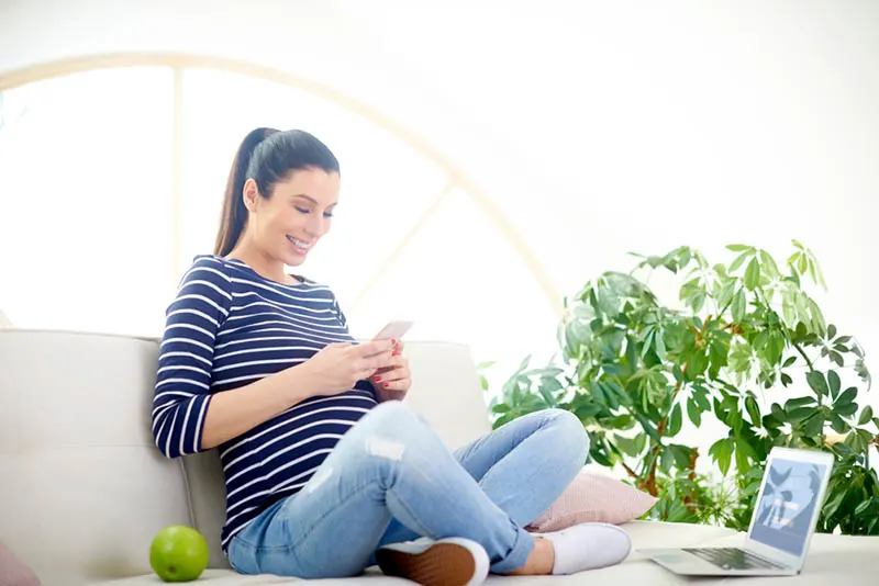 lächelnde schwangere Frau sitzt mit einem Apfel auf der Couch und schreibt eine SMS