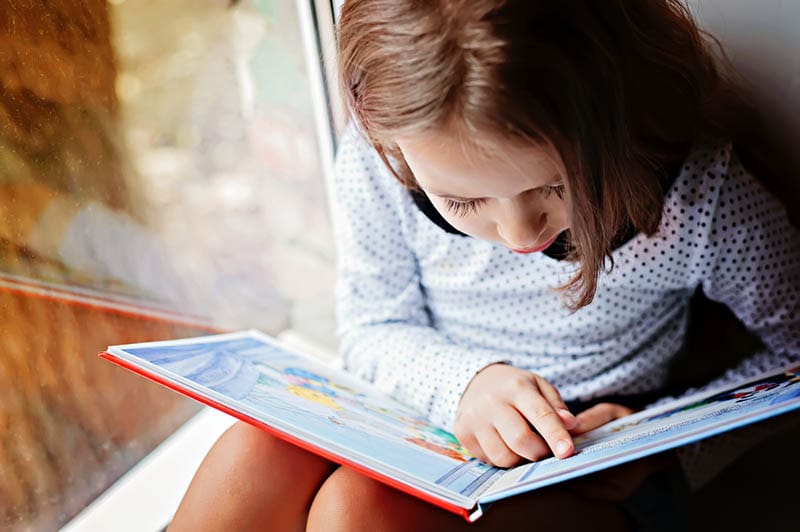 niedlichen kleinen Mädchen lesen ein Buch in der Nähe des Fensters
