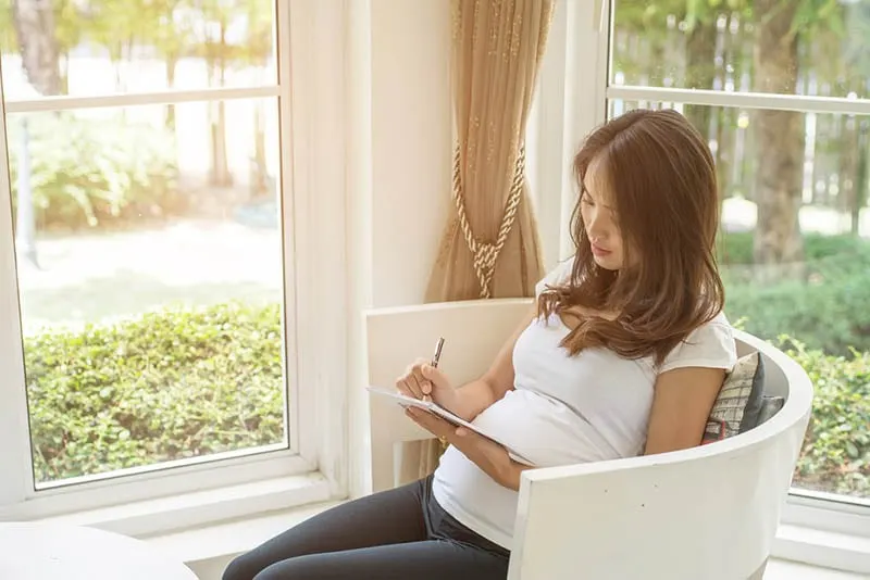 schöne schwangere Frau schreibt eine Karte und sitzt im Stuhl