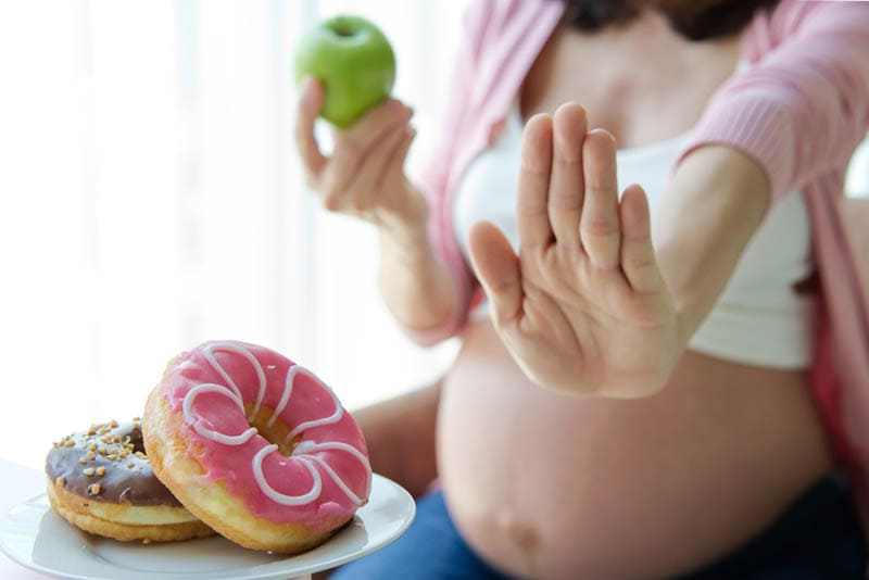 schwangere Frau hält einen Apfel und lehnt Donuts auf Teller ab