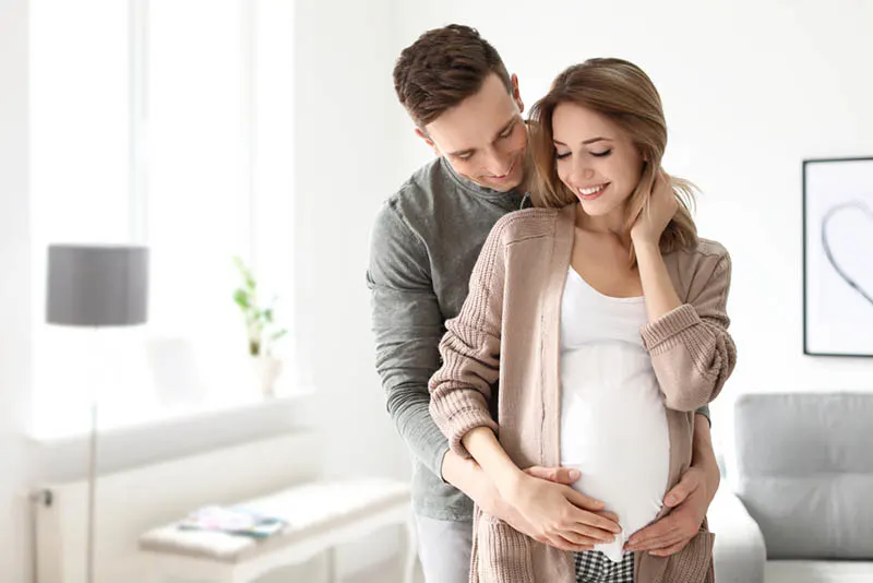 schwangere Frau steht mit ihrem Mann und beide halten die Hände auf den Bauch zu Hause