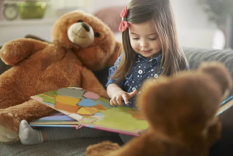 süßes Baby-Mädchen sitzt auf der Couch mit einem riesigen Teddybär und einem Buch im Arm