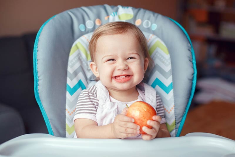 Süßes Baby Mädchen hält einen Apfel im Hochstuhl