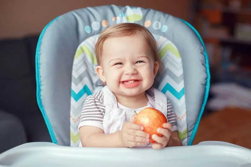 Süßes Baby Mädchen hält einen Apfel im Hochstuhl