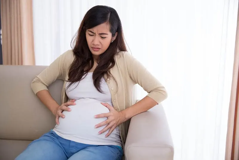 junge schwangere Frau sitzt in Schmerzen auf der Couch halten für Bauch