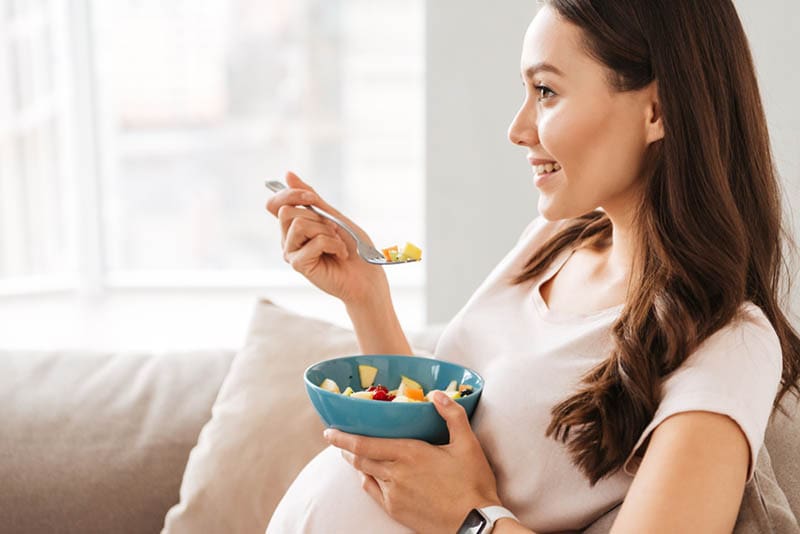 lächelnde schwangere Frau isst gesundes Essen auf dem Sofa