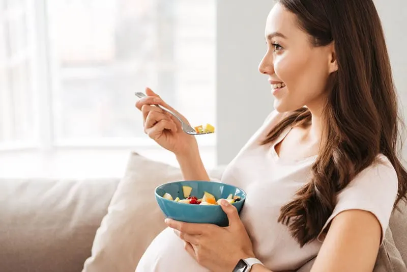 lächelnde schwangere Frau isst gesundes Essen auf dem Sofa
