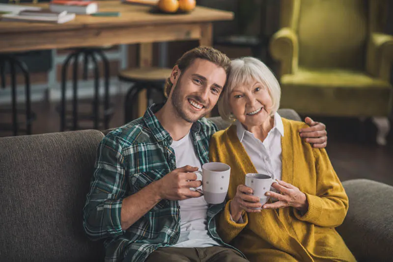 lächelnder Mann sitzt mit älterer Frau und hält Tassen mit Kaffee