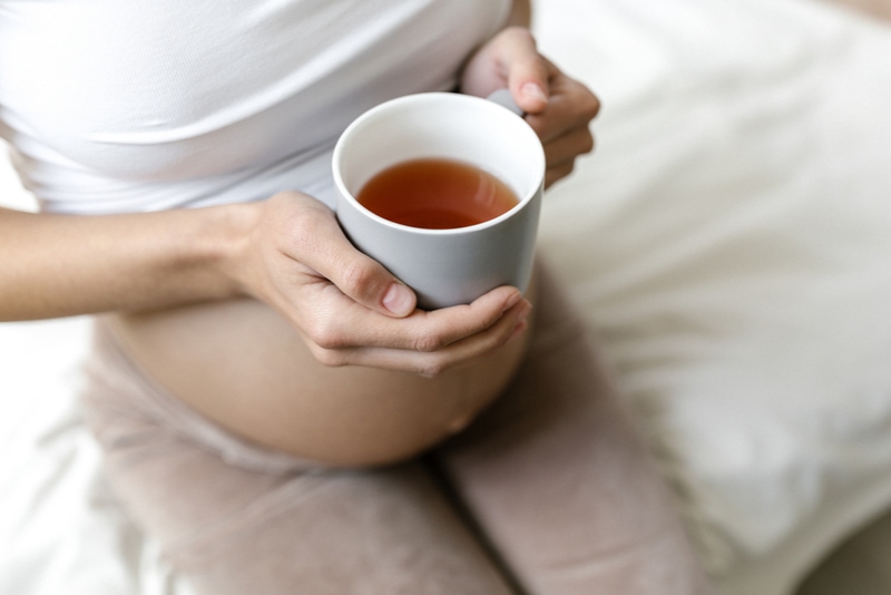 schwangere Frau sitzt auf dem Bett und hält eine Tasse Tee
