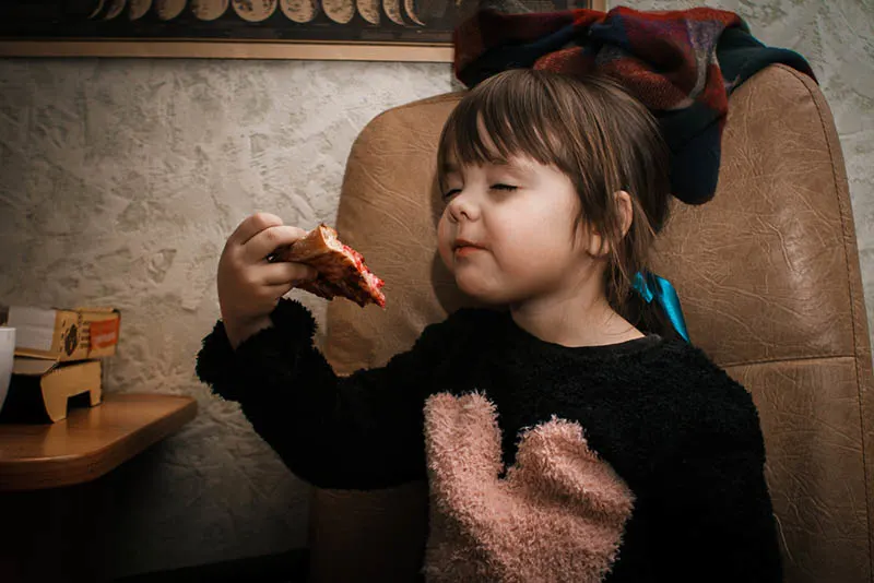 süßes kleines Mädchen mit Schleife auf dem Kopf isst ein Pizzastück