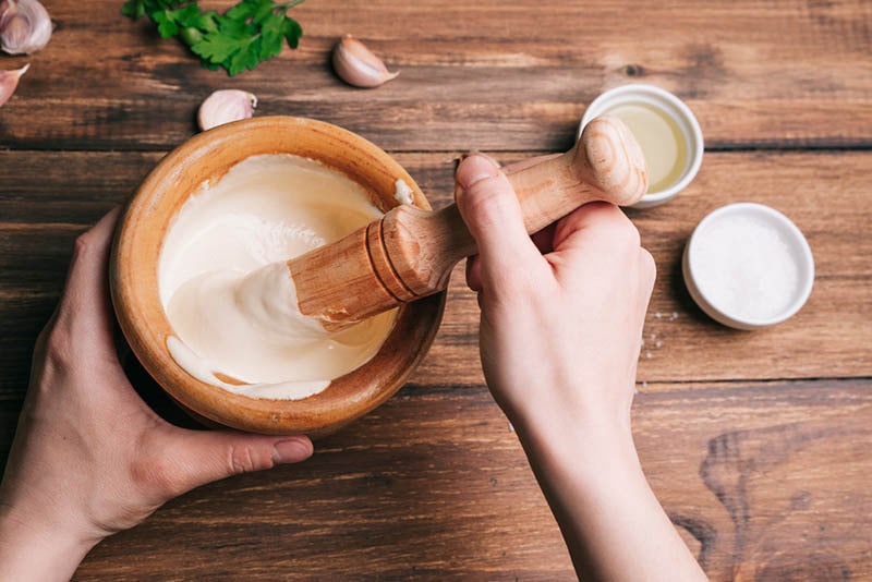Hände rühren Aioli-Sauce in einem Holzmörser auf einem Holztisch