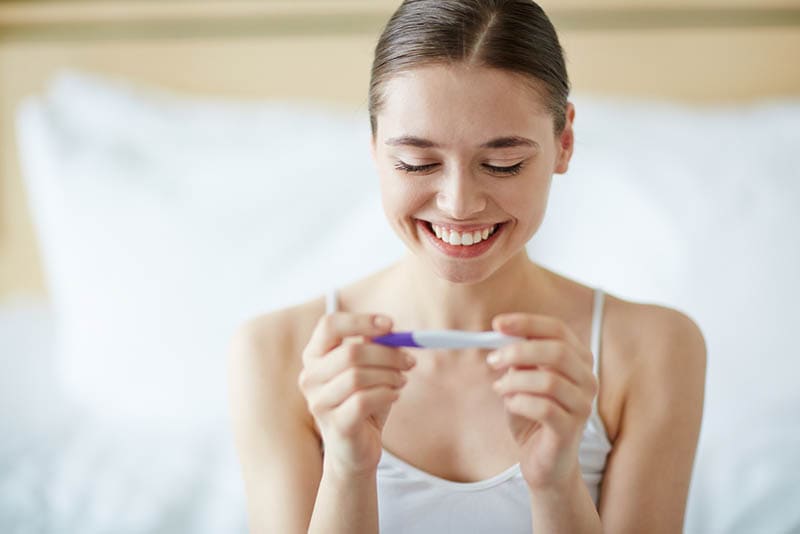 Junge glückliche Frau schaut auf Schwangerschaftstest im Schlafzimmer