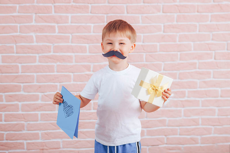 Kleiner Junge mit Schnurrbart hält ein Geschenk in seinen Händen