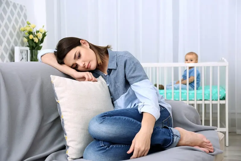 depressive Frau sitzt auf der Couch, während Baby in der Krippe ist