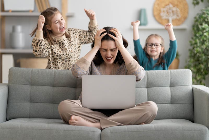 gestresste Mutter sitzt mit Laptop auf der Couch, während Kinder schreien