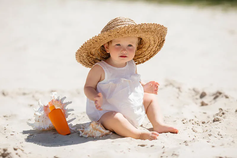 entzückendes kleines Mädchen mit Hut, der im Sand neben dem Sonnenschutz sitzt