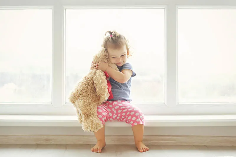 kleines Mädchen umarmt ihren Teddybär Spielzeug durch das Fenster