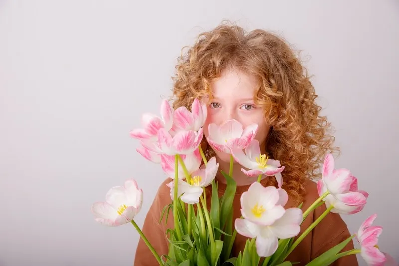 Mädchen mit lockigen Haaren hält sich rosa Blumen vor das Gesicht