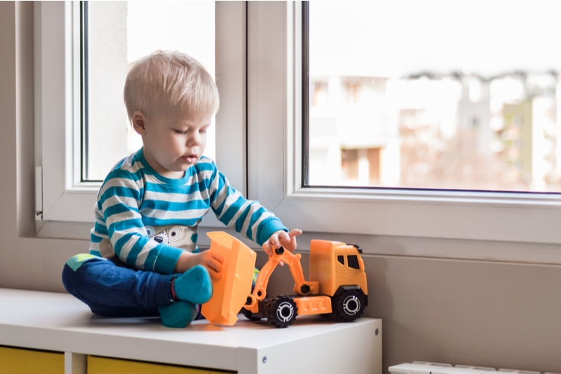 zweijähriger Junge sitzt am Fenster und spielt mit Spielzeuglaster