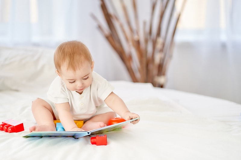 Baby spielt mit einem Buch, während es auf dem Bett sitzt
