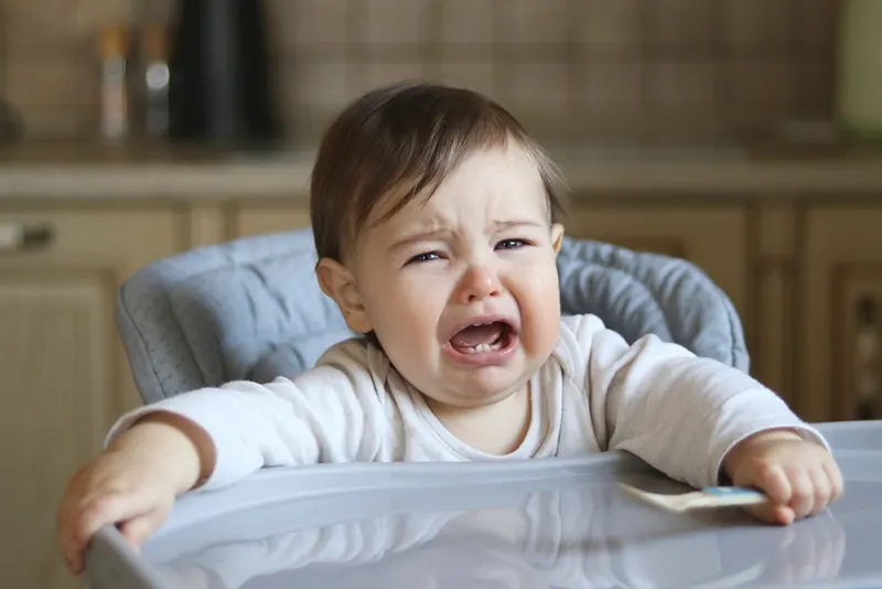 Baby weint im hohen Fütterstuhl sitzend und hält Löffel