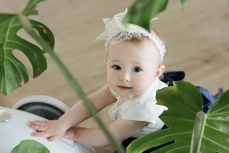 Babymädchen mit weißem Band auf dem Kopf, das neben Pflanzen im Haus steht