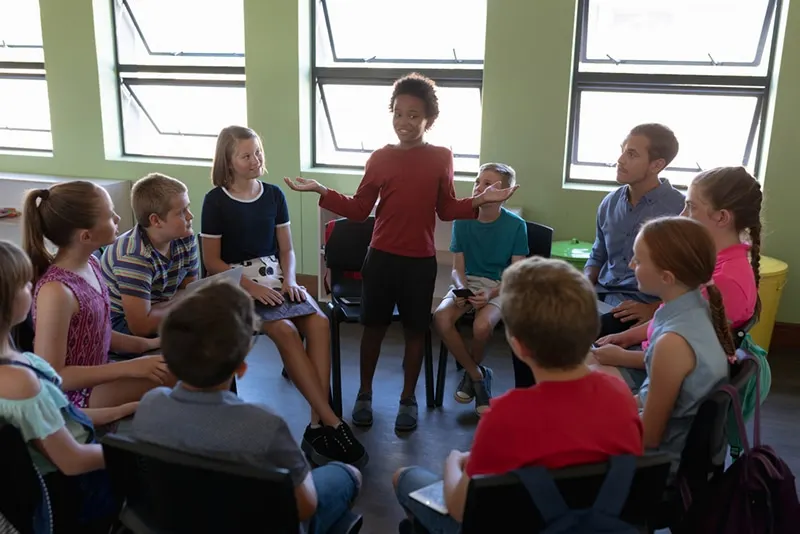 Gruppe von Grundschulkindern, die auf Stühlen im Kreis sitzen, während ein Mädchen steht und spricht