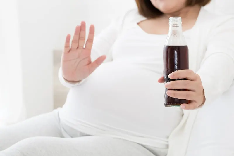 Schwangere Frau, die eine Flasche Cola hält und auf der Couch sitzt