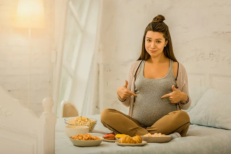 Schwangere Frau isst verschiedenes Essen auf dem Bett sitzend